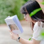 A young woman sits outdoors, absorbed in reading a book during a sunny summer day.